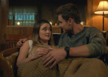 "A young couple sits close together on a chair, wrapped in a blanket in a cozy, warmly lit wooden room with a lamp, shelves, and candles in the background, creating an intimate and comfortable atmosphere."