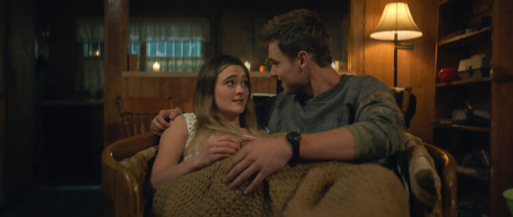 "A young couple sits close together on a chair, wrapped in a blanket in a cozy, warmly lit wooden room with a lamp, shelves, and candles in the background, creating an intimate and comfortable atmosphere."