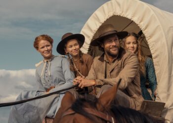"A man and three women in pioneer-era clothing ride a horse-drawn covered wagon outdoors, under a blue sky with clouds, suggesting a journey or travel in a historical rural setting."