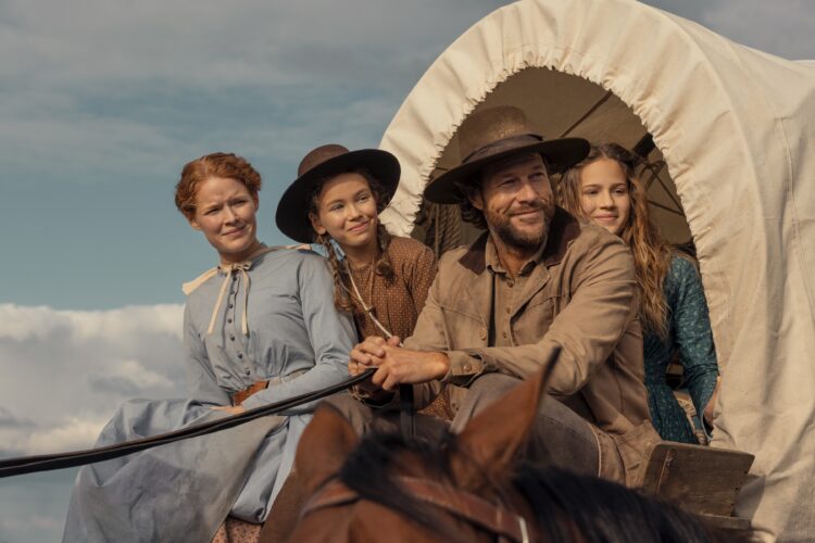 "A man and three women in pioneer-era clothing ride a horse-drawn covered wagon outdoors, under a blue sky with clouds, suggesting a journey or travel in a historical rural setting."