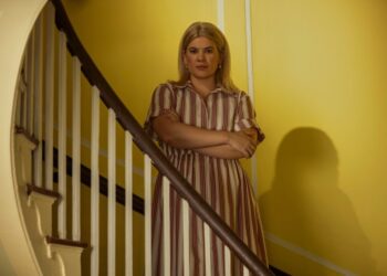 "A blonde woman in a striped dress stands with arms crossed by a wooden staircase inside a house with yellow walls, looking directly at the camera in soft indoor lighting."