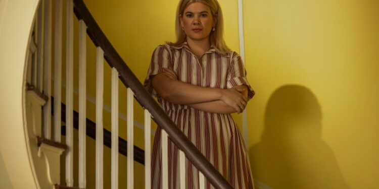 "A blonde woman in a striped dress stands with arms crossed by a wooden staircase inside a house with yellow walls, looking directly at the camera in soft indoor lighting."