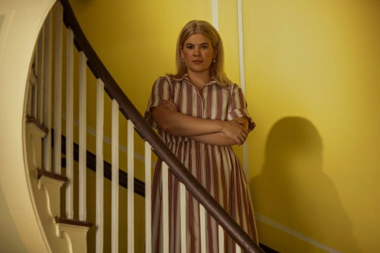 "A blonde woman in a striped dress stands with arms crossed by a wooden staircase inside a house with yellow walls, looking directly at the camera in soft indoor lighting."
